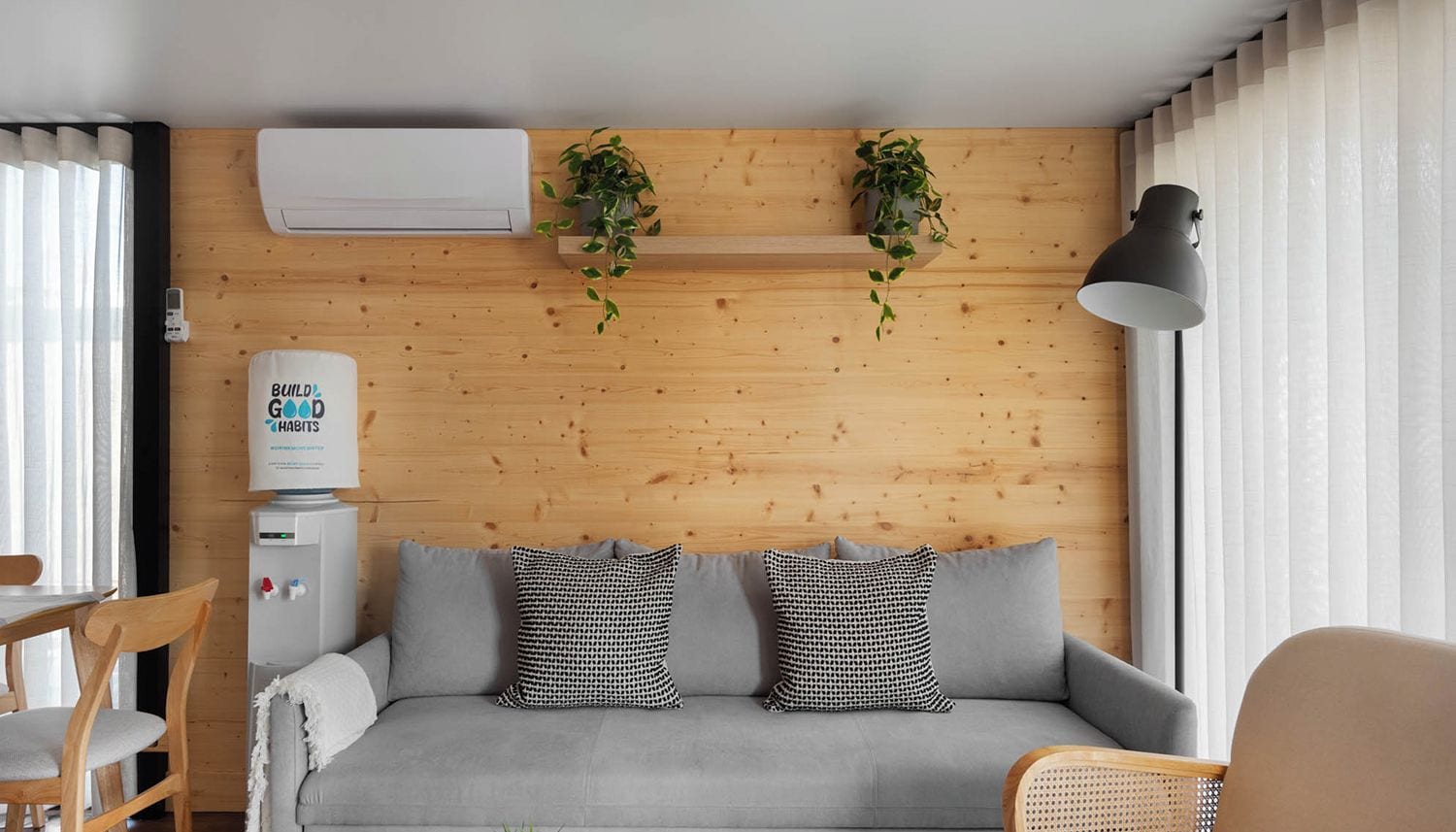 A modern living room with a grey sofa, two patterned cushions, a wooden wall, a water dispenser, and a potted plant.