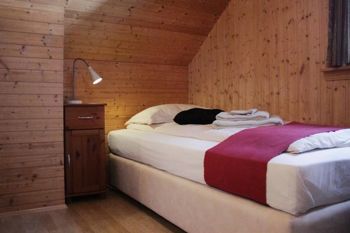 Una cama individual con ropa de cama blanca y una manta roja, una mesita de noche de madera con una lámpara, y un panel de madera.
