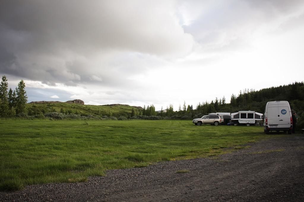 A campground with tents and vehicles, surrounded by green fields and hills under a cloudy sky.