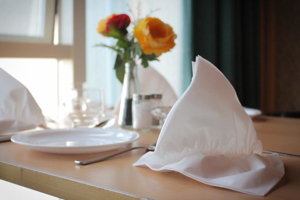 A neatly set dining table with white plates, a folded napkin, and a vase with yellow roses in the background.