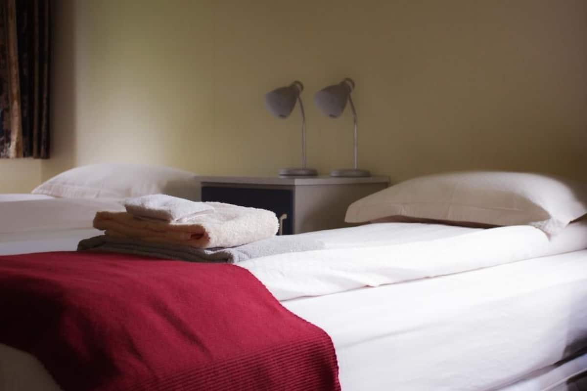 Habitación de hotel con dos camas, una colcha roja sobre una de ellas y una pila de toallas dobladas en la mesita de noche.