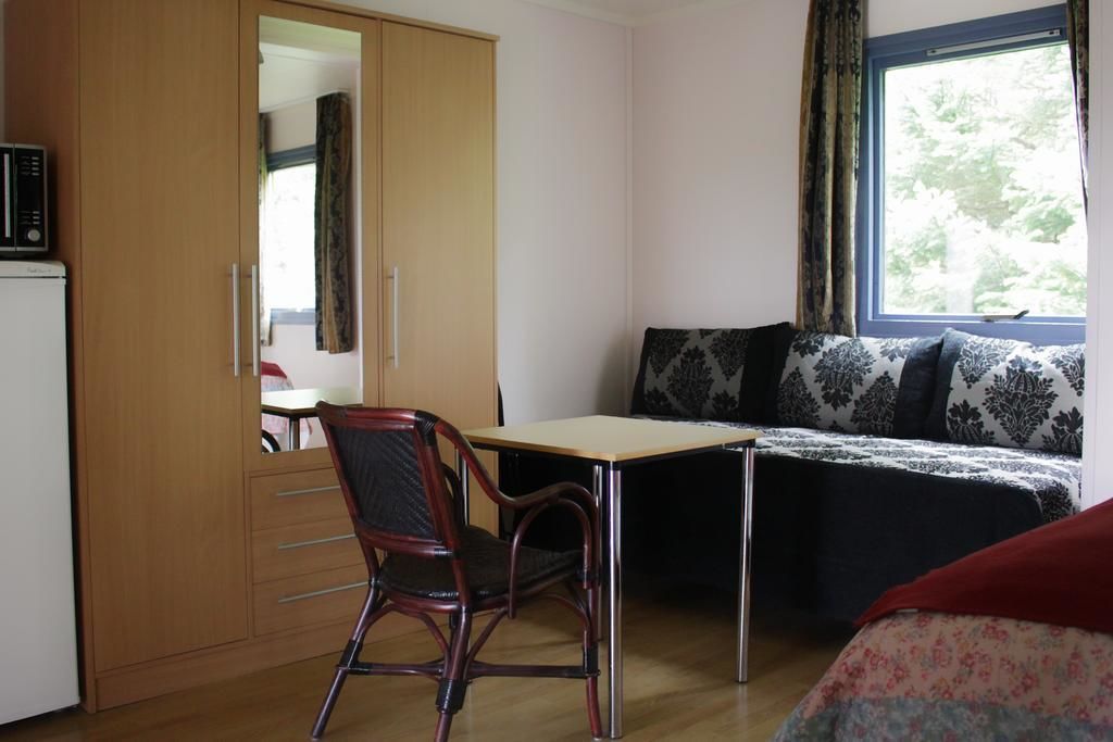 A room with a wooden wardrobe, a desk, a chair, a bed with patterned bedding, and a window with curtains.