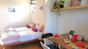 A small, cozy room with a bed, a desk, and a chair. There are pillows on the bed, a plant on the desk, and a small shelf above the desk. - Bnbook Passirana Studio with fast wifi (RHO)