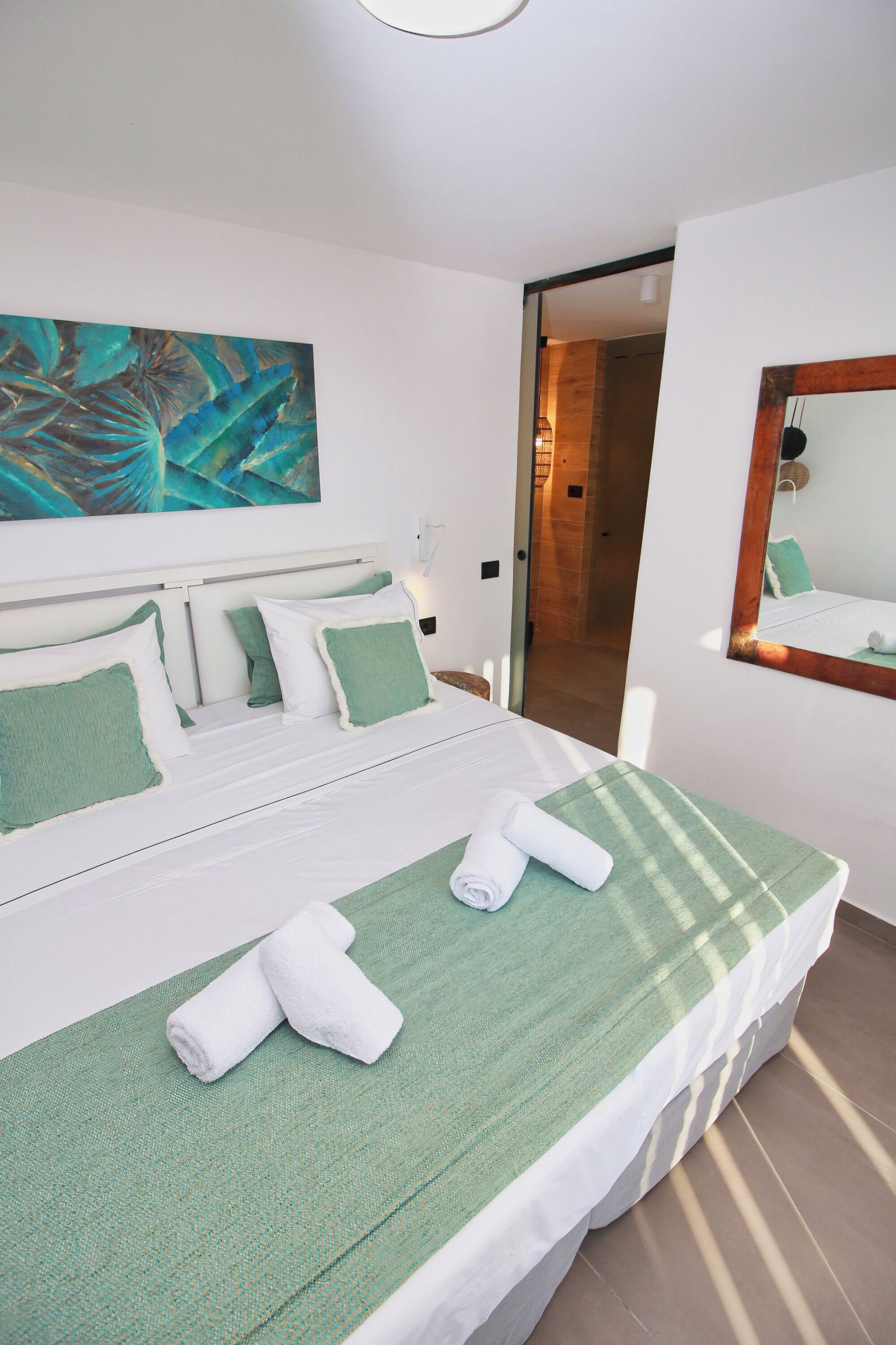 Una cama bien tendida con ropa de cama verde y dos toallas enrolladas, un cuadro abstracto grande en la pared y un espejo que refleja parte de la habitación.