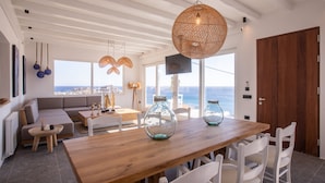 A modern living room with a wooden dining table, white chairs, and a large window overlooking the sea.