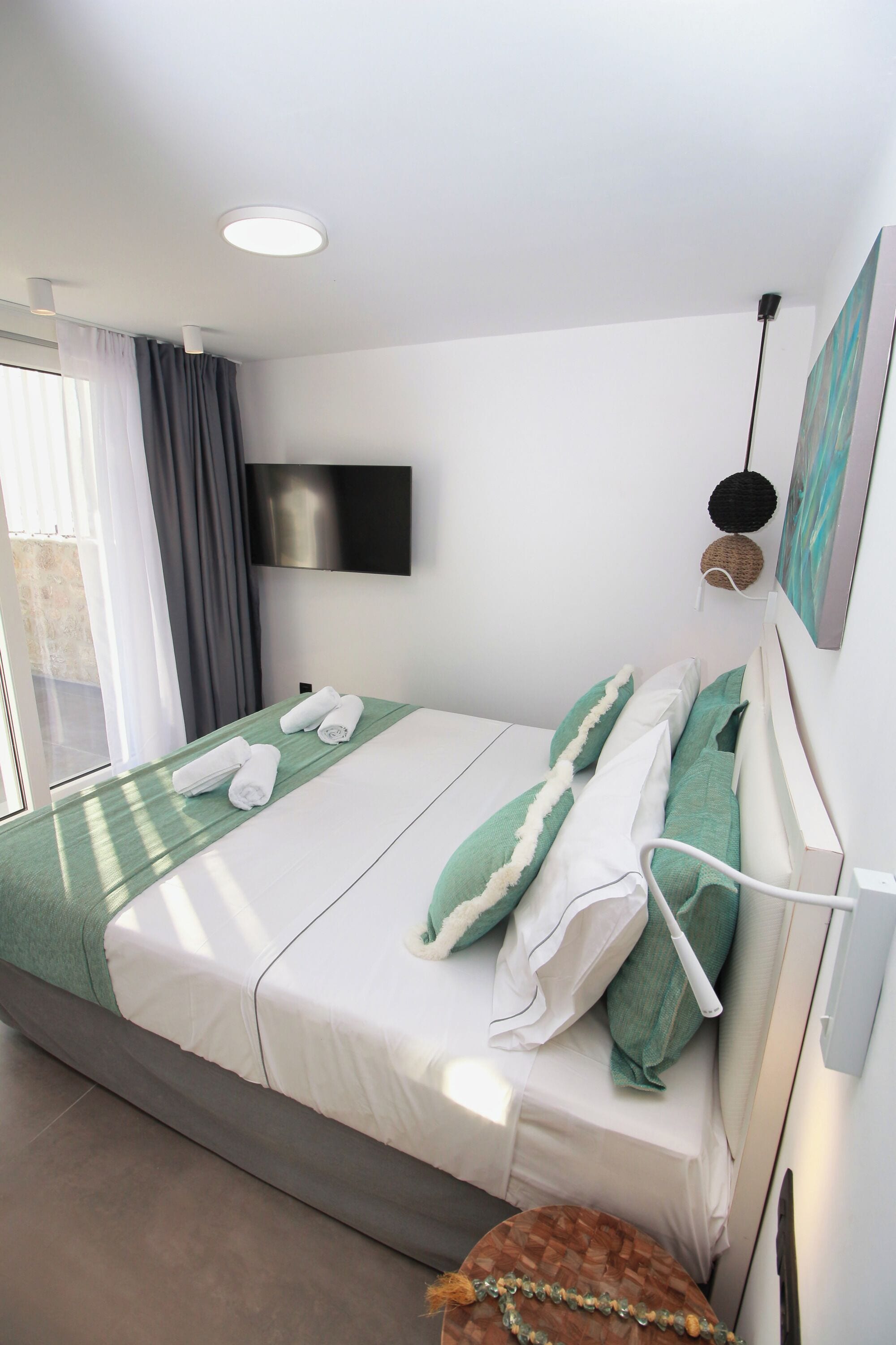 Una cama bien hecha con ropa de cama blanca y verde, un televisor de pantalla plana en la pared y una ventana con cortinas.