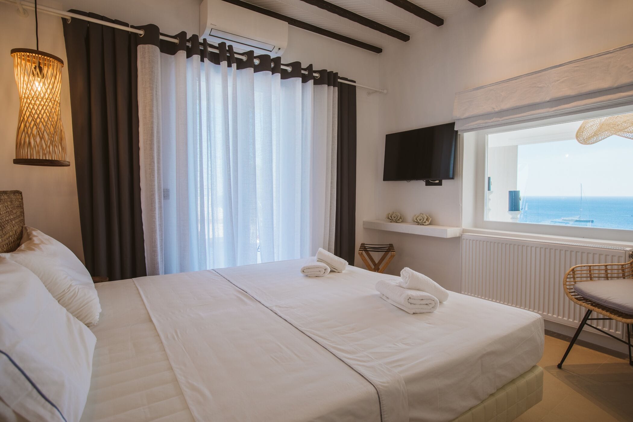 A hotel room with a bed, a TV, a window with a view of the sea, and a wicker chair.