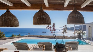 A poolside area with wicker pendant lights, a view of the sea, and a table with a bowl of fruit.
