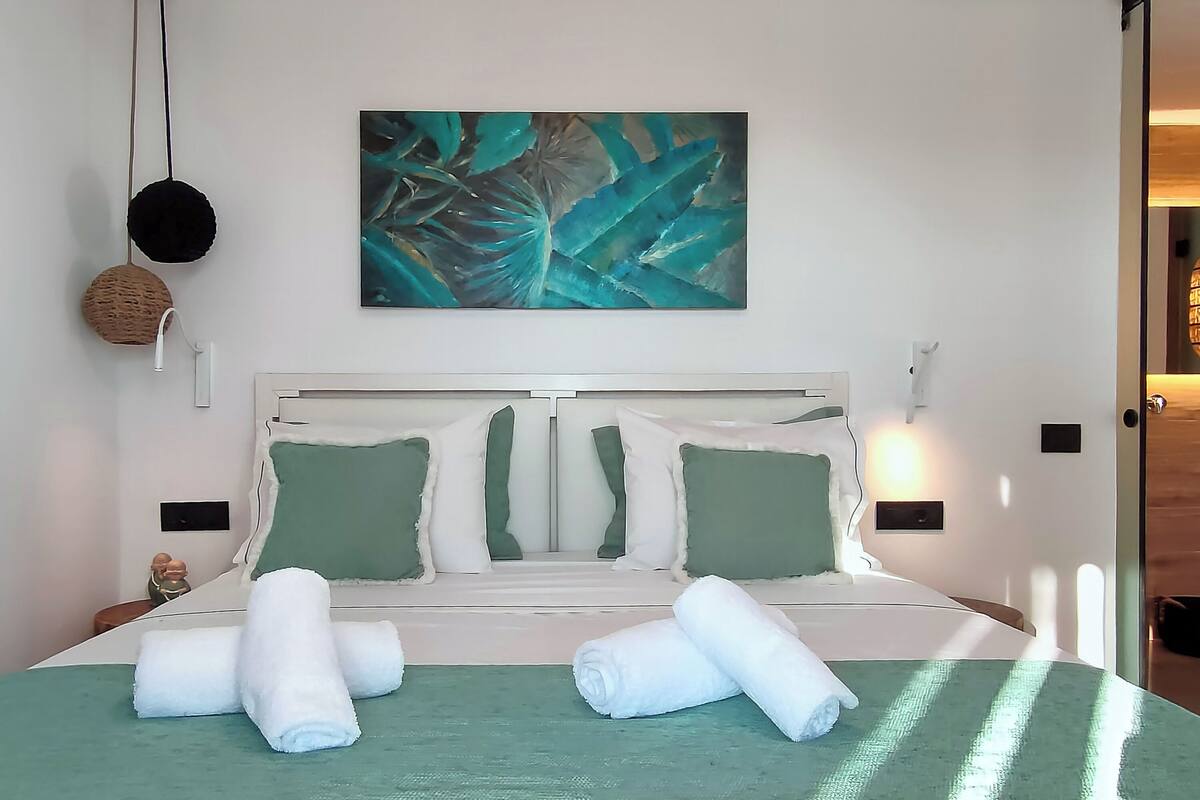 Una cama bien hecha con ropa de cama verde y almohadas blancas, un cuadro sobre ella y una lámpara de pared.