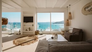 A modern living room with a large window overlooking the sea, a flat-screen TV mounted on the wall, and a cozy seating area with a brown sofa and a coffee table.