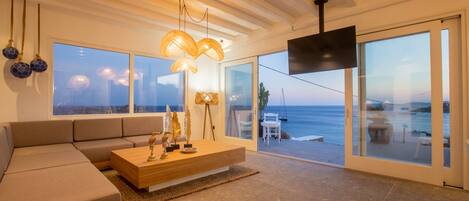 A modern living room with a large sofa, a wooden coffee table, and a view of the sea through large glass doors.