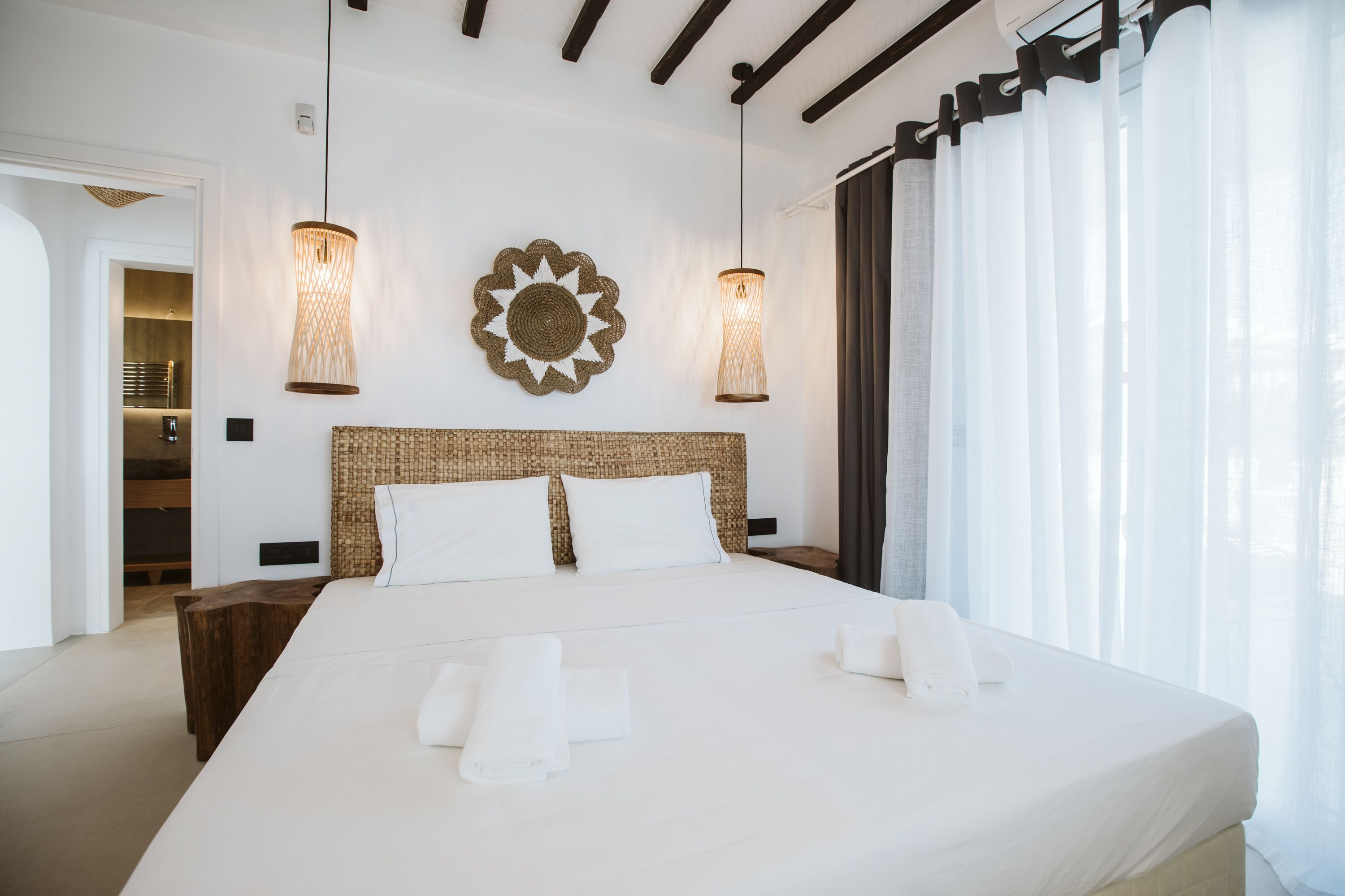 A modern hotel room with a large bed, two wall-mounted lamps, a woven headboard, and a view of the outdoors through sheer curtains.