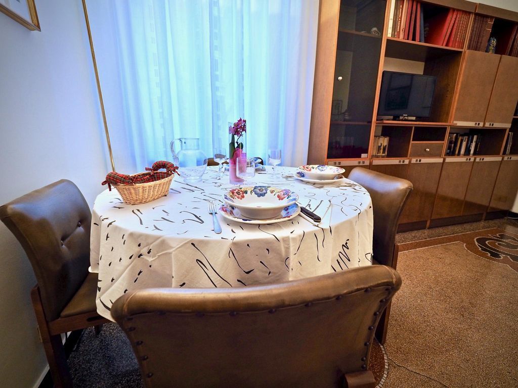 A dining area with a round table set for two, a wicker basket, a vase with flowers, and a television in the background.