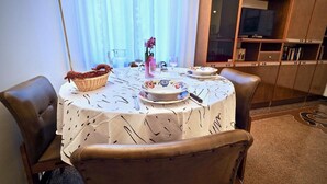 Un comedor con una mesa redonda preparada para dos personas, una cesta de mimbre, un florero con flores y un televisor al fondo.