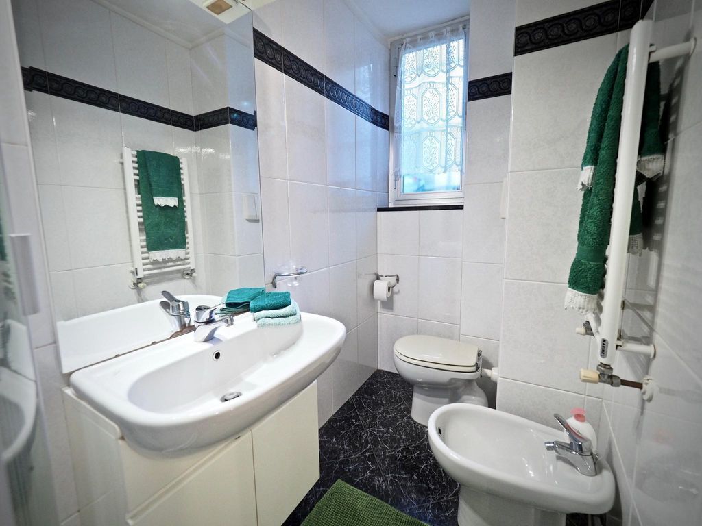 A bathroom with a white sink, a white toilet, and a bidet. There is a window with decorative glass and a green towel hanging on the wall.