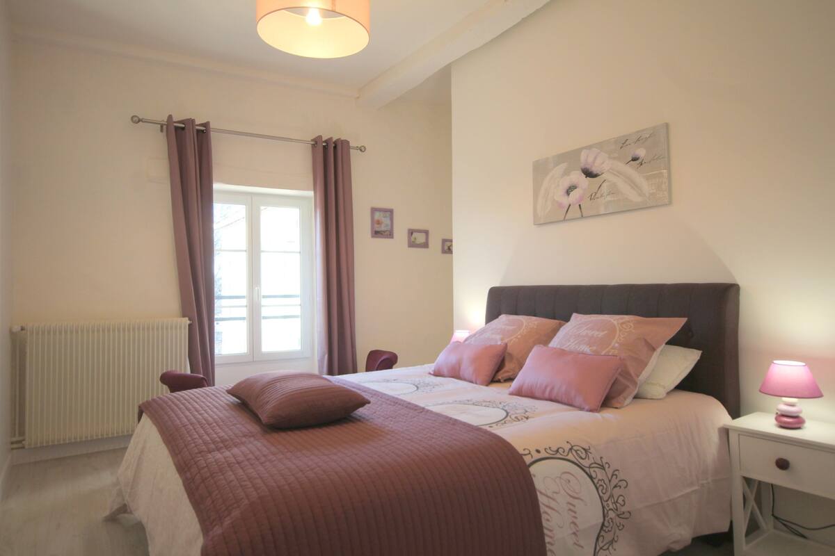 Un dormitorio con una cama, una mesita de noche, una lámpara, una ventana con cortinas y un cuadro con marcos.