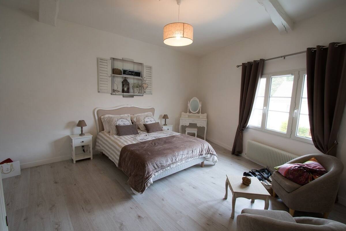 Un dormitorio con cama, mesitas de noche, cómoda, silla, mesa, lámpara colgante y ventana con cortinas.