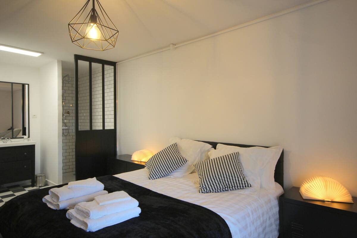 Un dormitorio moderno con una cama grande, ropa de cama blanco y negro, un cabecero negro, una mesita de noche con una lámpara y un espejo sobre el aparador.