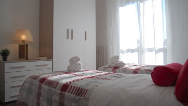 Une chambre d’hôtel avec deux lits, une grande armoire, une table de chevet avec une lampe et une fenêtre avec des rideaux légers.