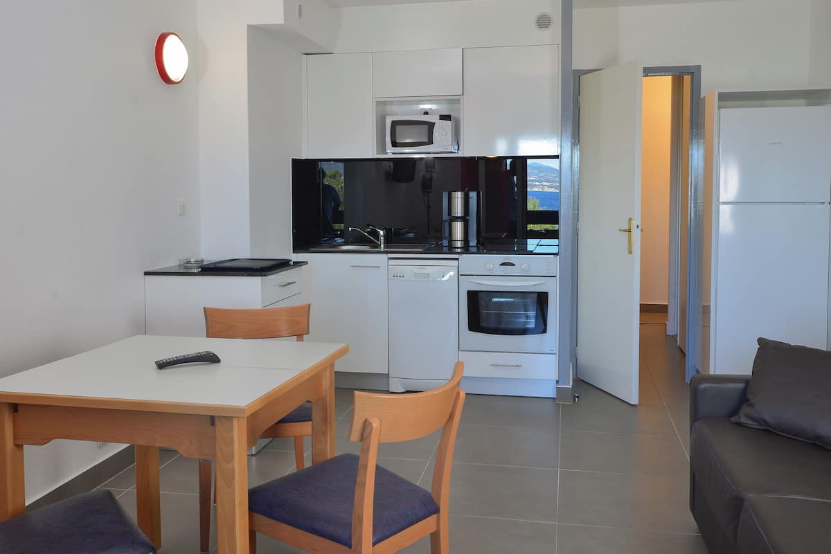 A compact kitchen with white cabinets, a microwave, a stove, and a dishwasher. There is a dining table with chairs and a sofa in the living area.