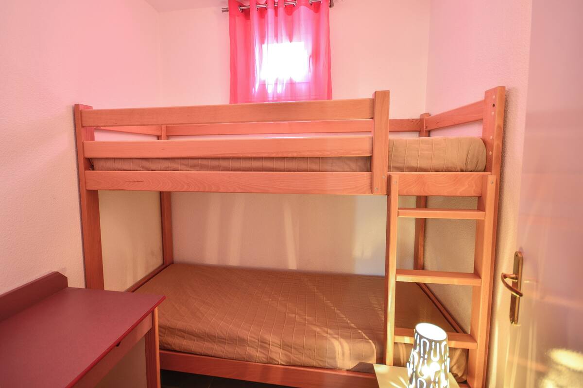 A bunk bed with a wooden frame, a small desk, and a window with pink curtains.