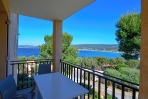 Un balcón con mesa y sillas, con vista al mar, árboles y cielo despejado.