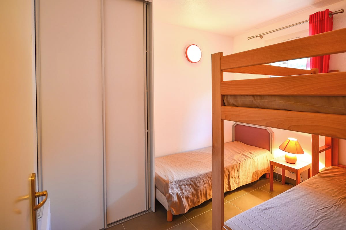A dormitory room with bunk beds, a bedside table with a lamp, and a window with red curtains.