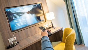 A person holding a remote control in front of a flat-screen TV mounted on a wooden wall, with a lamp and a glass on a small table below. - Hotel Meyerink (Vreden)