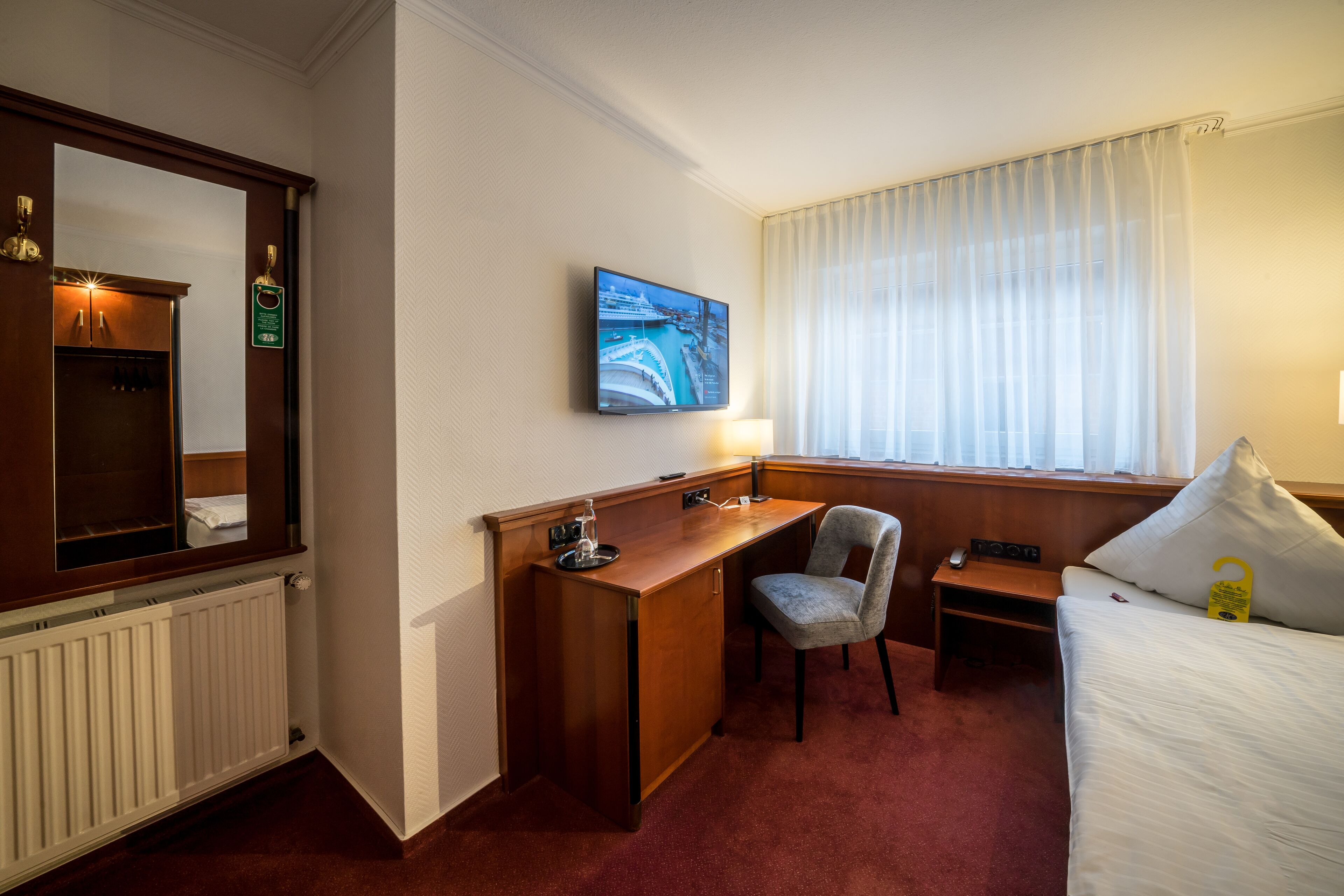 Quarto de hotel com escrivaninha de madeira, uma cadeira, uma cama, televisão e janela com cortinas.