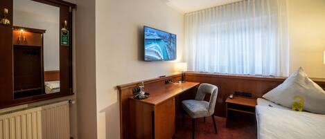 Quarto de hotel com escrivaninha de madeira, uma cadeira, uma cama, televisão e janela com cortinas.