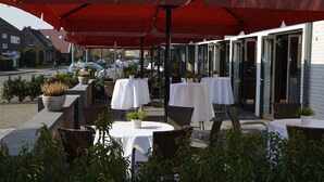 An outdoor patio with tables and chairs, covered by a large red umbrella, and potted plants. - Hotel Meyerink (Vreden)