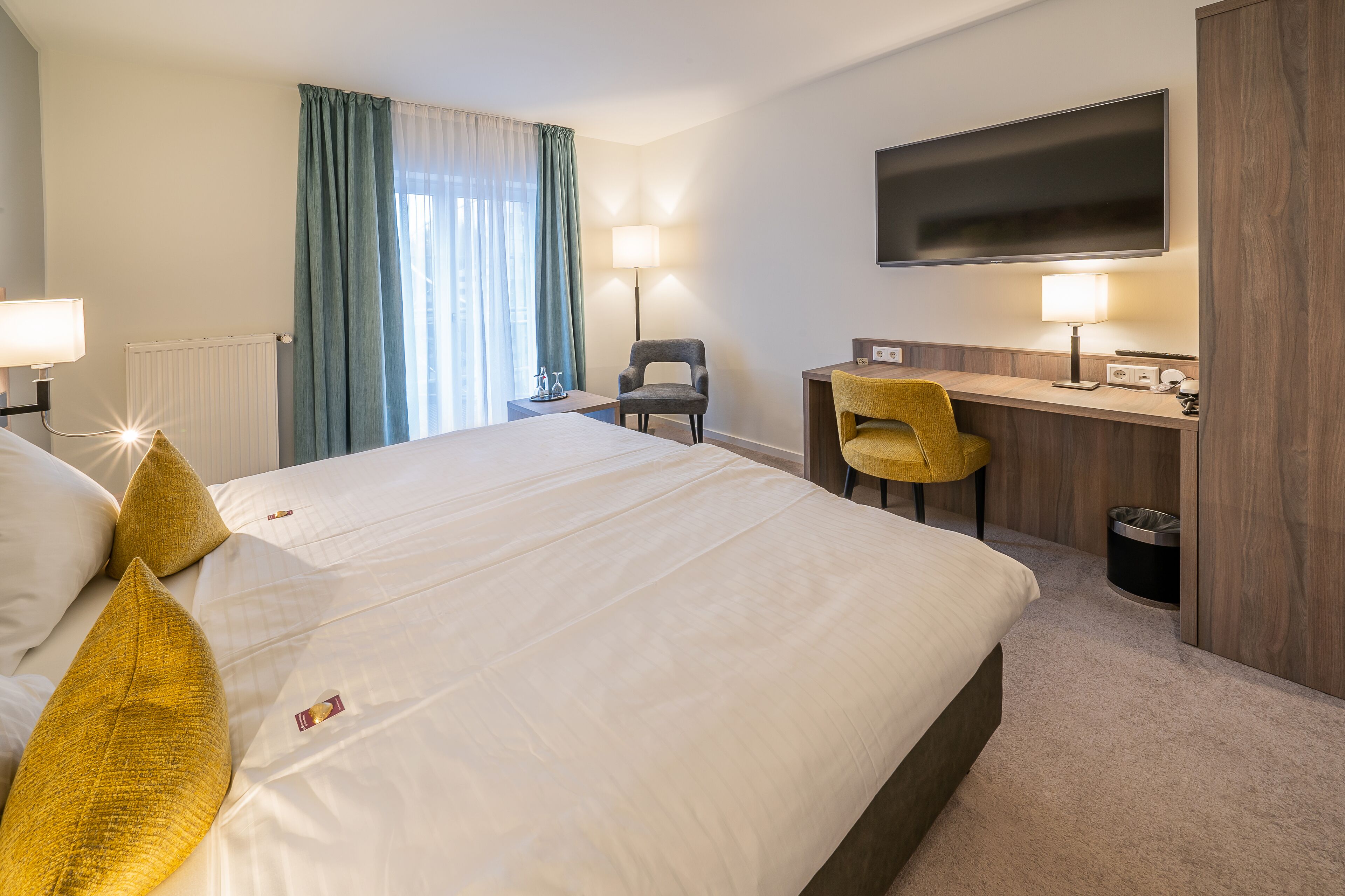 A hotel room with a large bed, a desk, a chair, a TV, and a lamp.