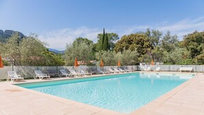 A rectangular swimming pool with clear blue water, surrounded by white lounge chairs and orange umbrellas, set against a backdrop of trees and mountains. - Village Vacances La Fontaine d'Annibal (Buis-les-Baronnies)