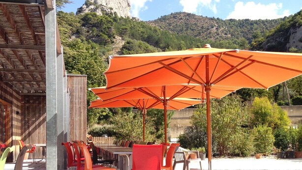An outdoor patio with tables and chairs, orange umbrellas, and a mountainous backdrop. - Village Vacances La Fontaine d'Annibal (Buis-les-Baronnies)
