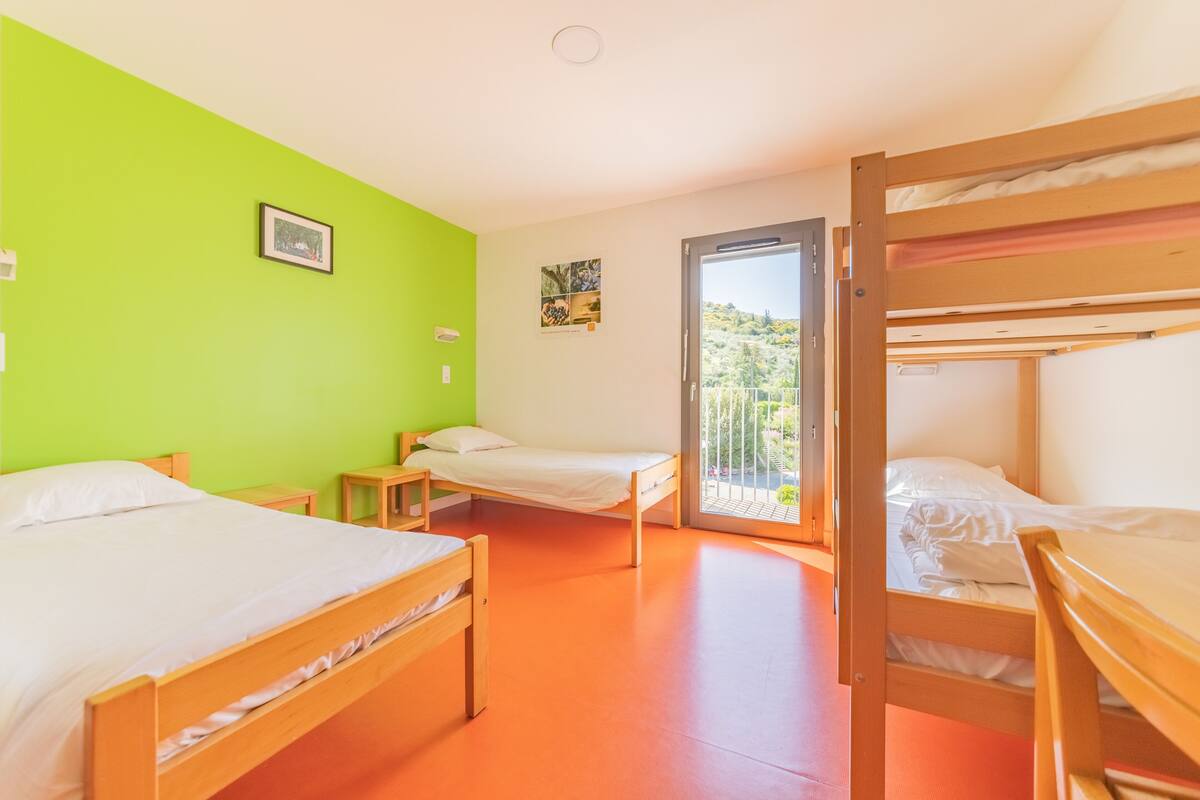 Ein Zimmer mit zwei Einzelbetten, einem Holzbettgestell, einem kleinen Tisch und einem Stuhl. Eine Tür führt zu einem Balkon mit Blick auf Grünflächen.