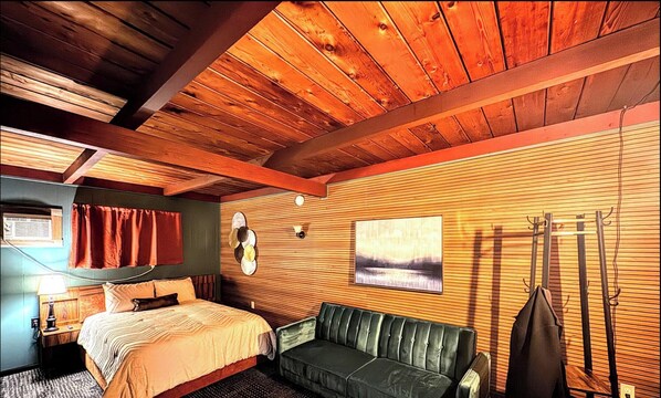 A room with a bed, a green sofa, wooden paneling, a painting, and a coat rack. - adoba® inn & suites (Charlevoix)