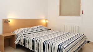 A single bed with a striped bedspread, a bedside table, a wall-mounted light, and a window with blinds. - VVF Résidence Arcachon (Arcachon)