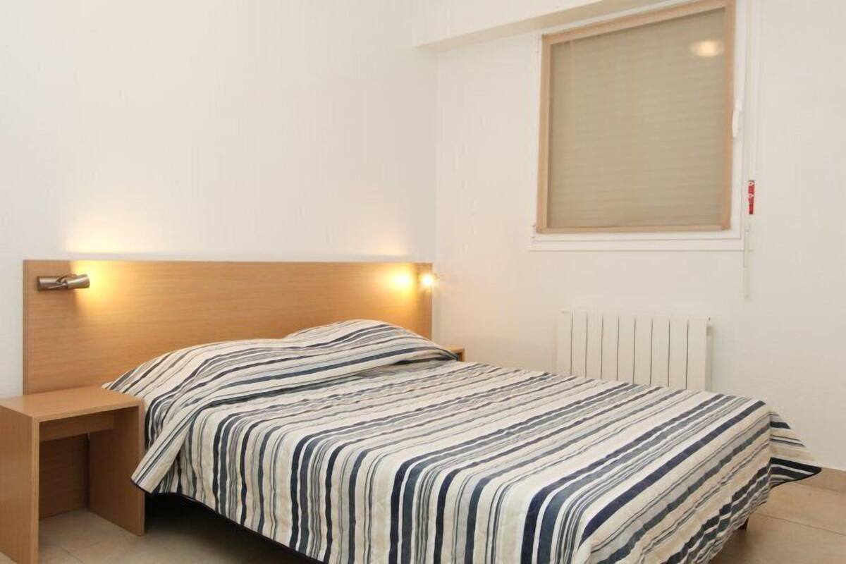 A single bed with a striped bedspread, a bedside table, a wall-mounted light, and a window with blinds.