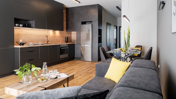 Eine moderne Küche mit dunklen Schränken, einer Holz Arbeitsplatte und einem eingebauten Kühlschrank. Ein Essbereich mit Tisch und Stühlen lädt zum Verweilen ein, und ein graues Sofa mit einem gelben Kissen sorgt für Gemütlichkeit.