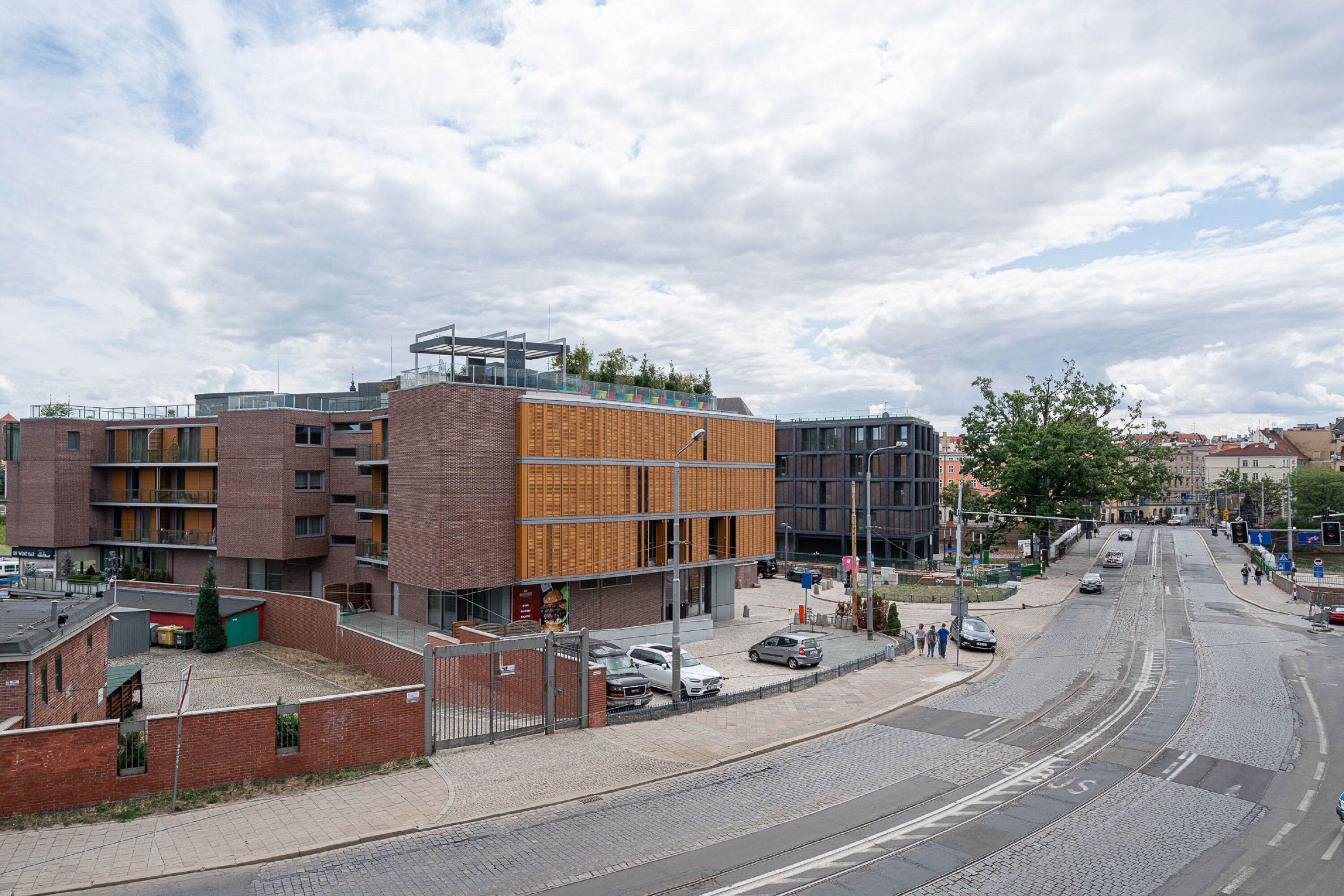A modern building with a mix of brick and wooden facades, a tram line, and a paved street with parked cars.