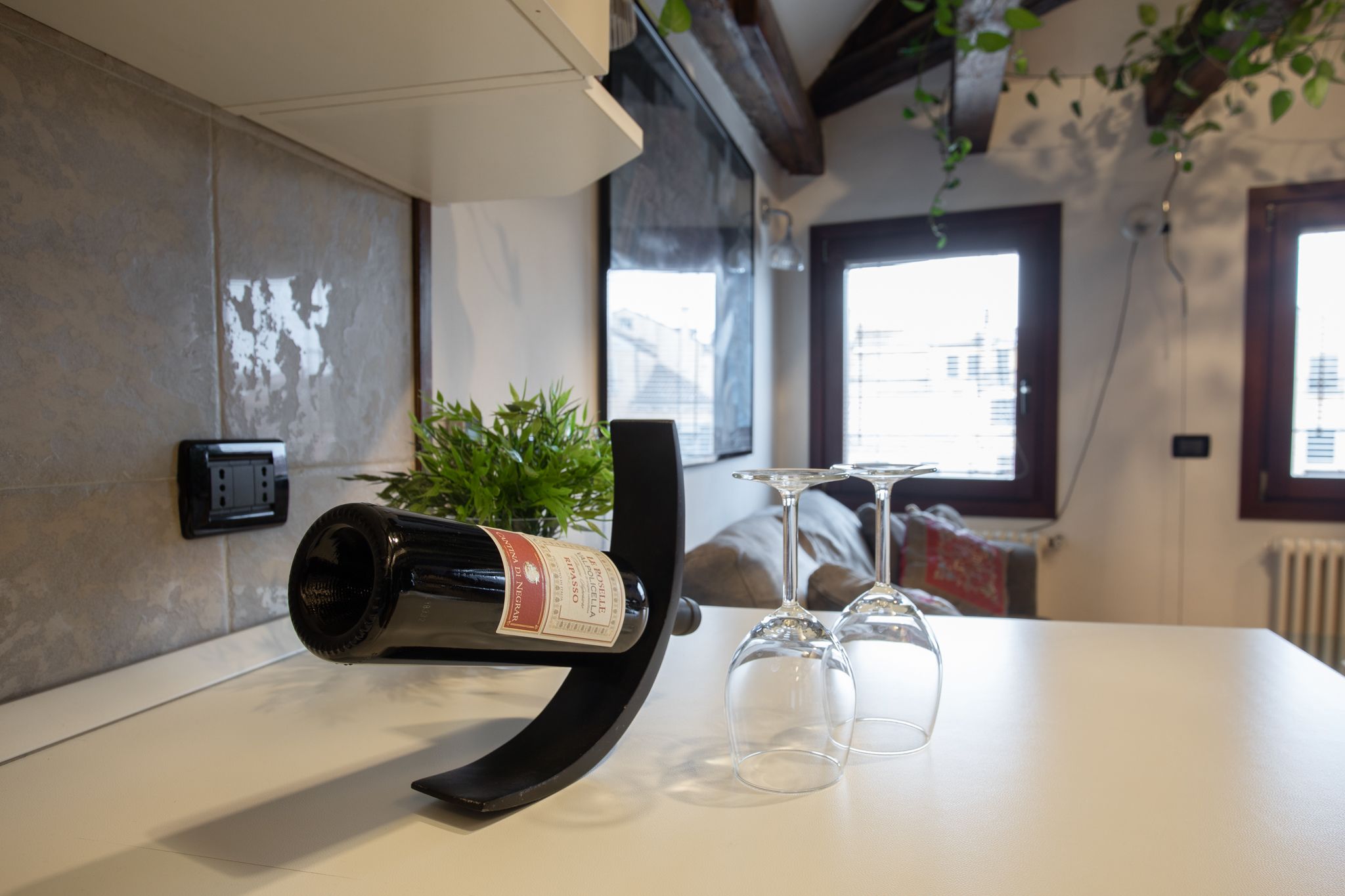 Una botella de vino sobre una mesa con dos copas de vino, una planta en maceta y una ventana con vista a edificios.