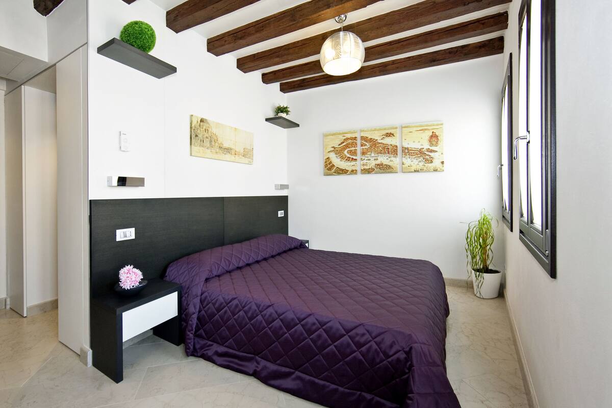 Un dormitorio con cama, mesita de noche, planta en maceta y obras de arte en las paredes.