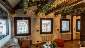 Un salon chaleureux avec un canapé, un pouf rouge, une table basse en verre et une vue sur les immeubles à travers de grandes fenêtres.