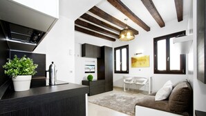 Une cuisine moderne avec des meubles de couleur sombre, un évier en acier inoxydable et une plante en pot. On y trouve un coin repas avec une table et des chaises blanches, ainsi qu’un canapé avec des coussins.