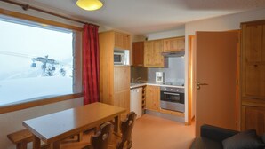 Une cuisine compacte dotée de meubles en bois, d’un four à micro-ondes, d’une machine à café et d’une cuisinière. On y trouve une table à manger avec des chaises, ainsi qu’un canapé avec un coussin.
