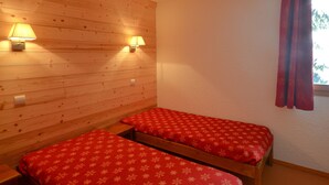 Une pièce aux murs recouverts de lambris en bois, avec deux lits dotés de draps rouges, deux lampes fixées au mur et une fenêtre ornée de rideaux rouges.