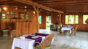 A rustic wooden interior with a dining area featuring tables and chairs, a bar area with bottles, and a view of greenery outside. - Esralina Pansiyon (Kemer)