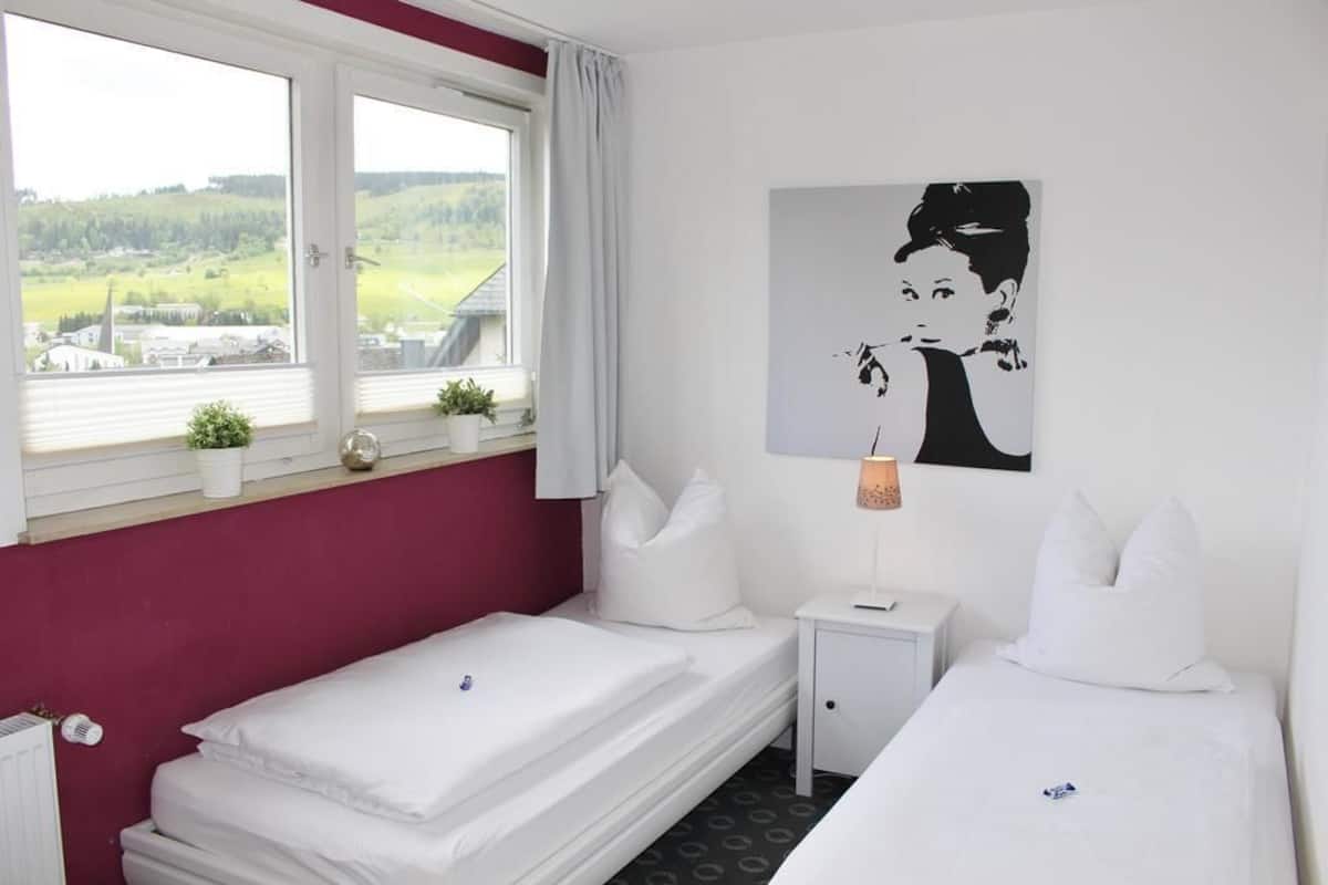 Ein Hotelzimmer mit zwei Betten, einem Fenster mit Blick auf die Landschaft, einem kleinen Nachttisch mit Lampe und einem an der Wand hängenden Kunstwerk.