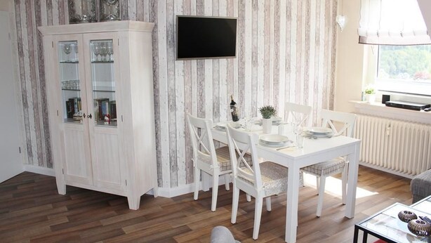 A dining area with a white table and chairs, a cabinet with glass doors, and a television mounted on the wall. - Löwenstein Ferienwohnungen (Willingen)