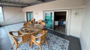Uma área de refeições com mesa de vidro redonda e cadeiras de vime, piso estampado, e vista para um quarto através de uma porta deslizante.